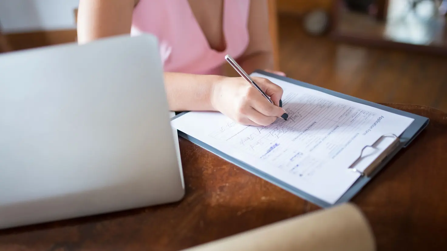 Une femme remplit un formulaire administratif sur un bureau en bois, l'ordinateur portable ouvert à côté pour faire ses démarches d'aides sociales en ligne