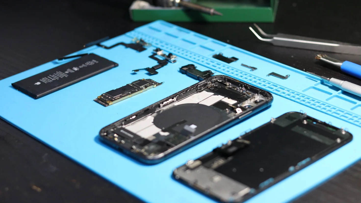 Smartphone entièrement démonté sur un tapis d'atelier bleu, batterie et carte mère posées à côté du châssis, outils de réparation autour