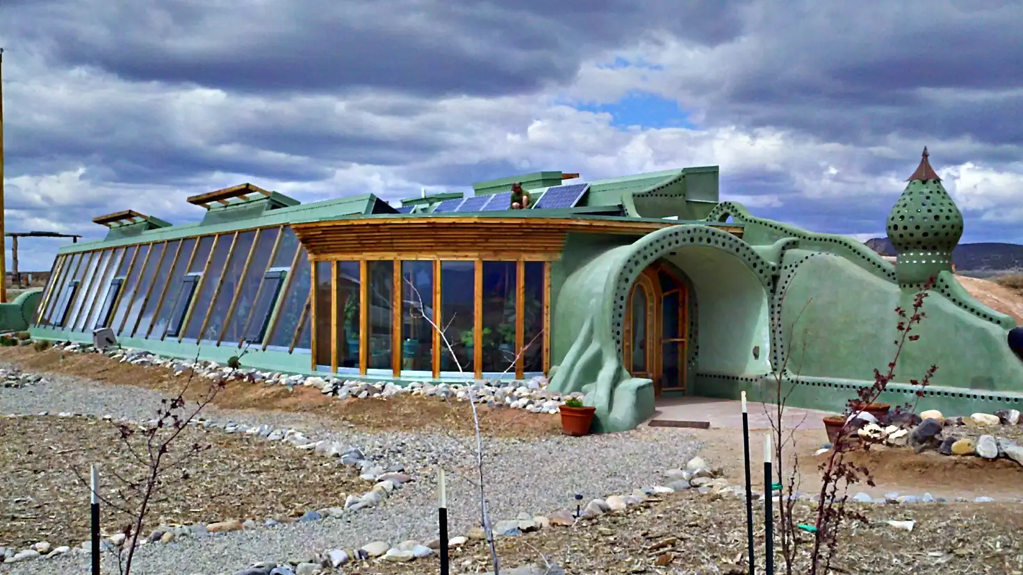 Earthship de la communauté Greater World à Taos : maison autonome aux murs incurvés en pneus et terre, bouteilles colorées encastrées et grande serre vitrée orientée plein sud