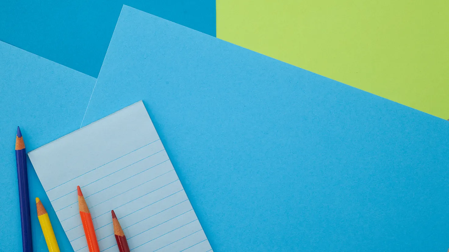 Crayons de couleur et bloc-notes ligné posés sur des feuilles colorées, pour préparer des fournitures scolaires pas cher