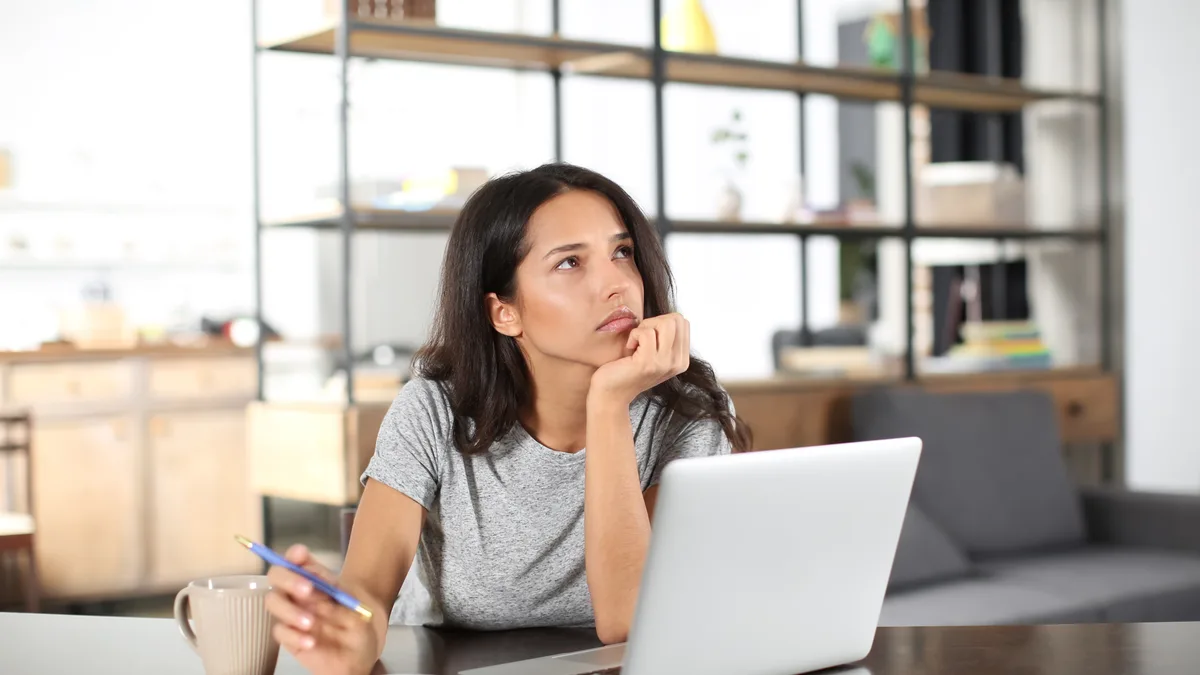 Idées reçues sur l'investissement : une jeune femme pensive, café et stylo en main, réfléchit à ses placements devant son ordinateur portable