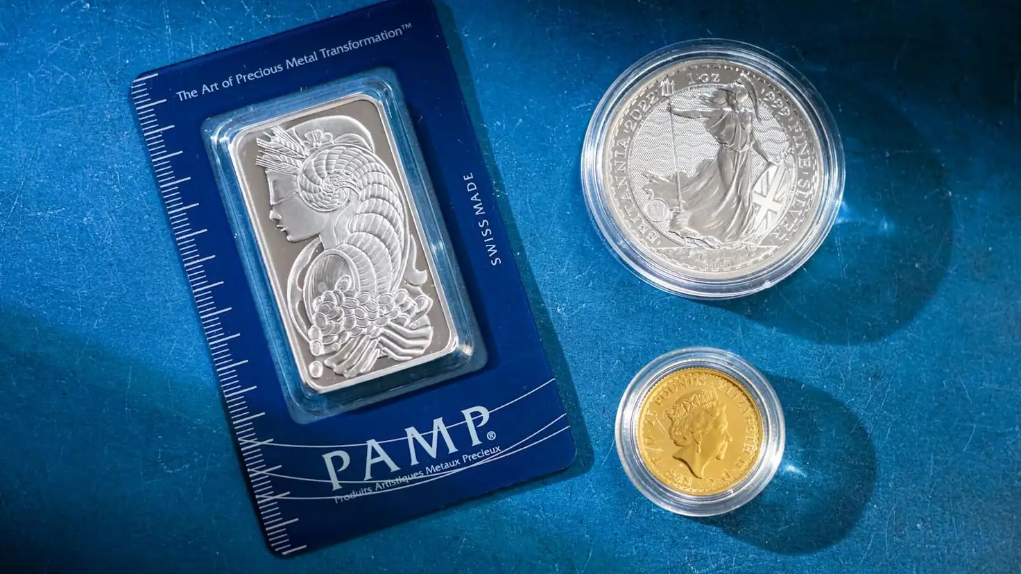 Investir dans l'argent métal : un lingot d'argent PAMP et une pièce d'argent Britannia posés sur fond bleu, à côté d'une petite pièce d'or, illustration de l'argent métal physique comme placement