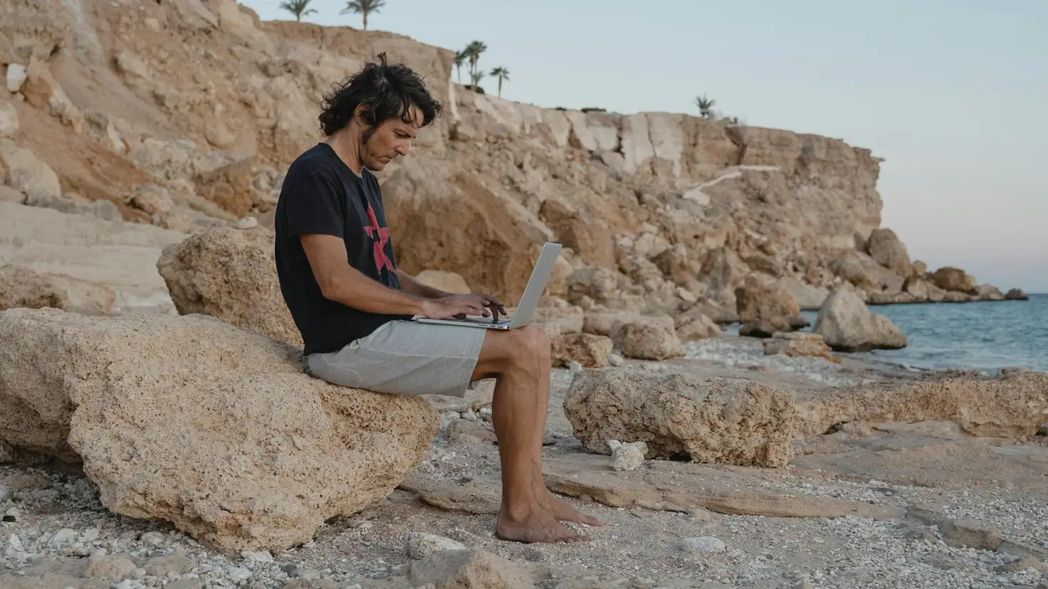 Mensonges sur l'indépendance financière : un homme travaille voûté sur son ordinateur portable, assis sur les rochers d'une plage, l'envers du décor vendu par le marketing de la liberté financière