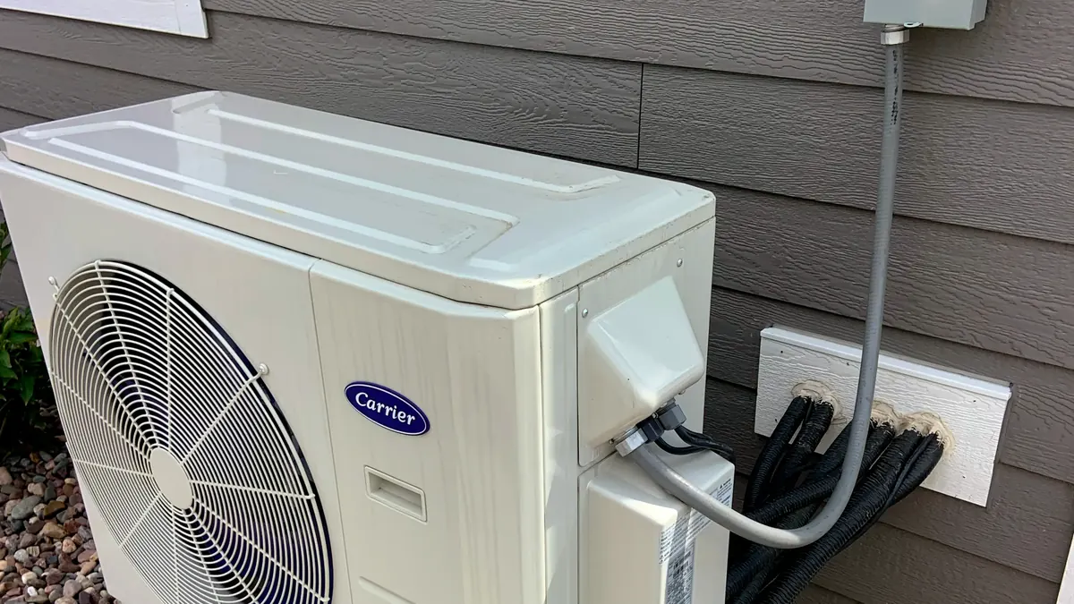 Unité extérieure de pompe à chaleur air-eau installée sur socle surélevé contre la façade d'une maison, avec ventilateur à grille et raccordements électriques