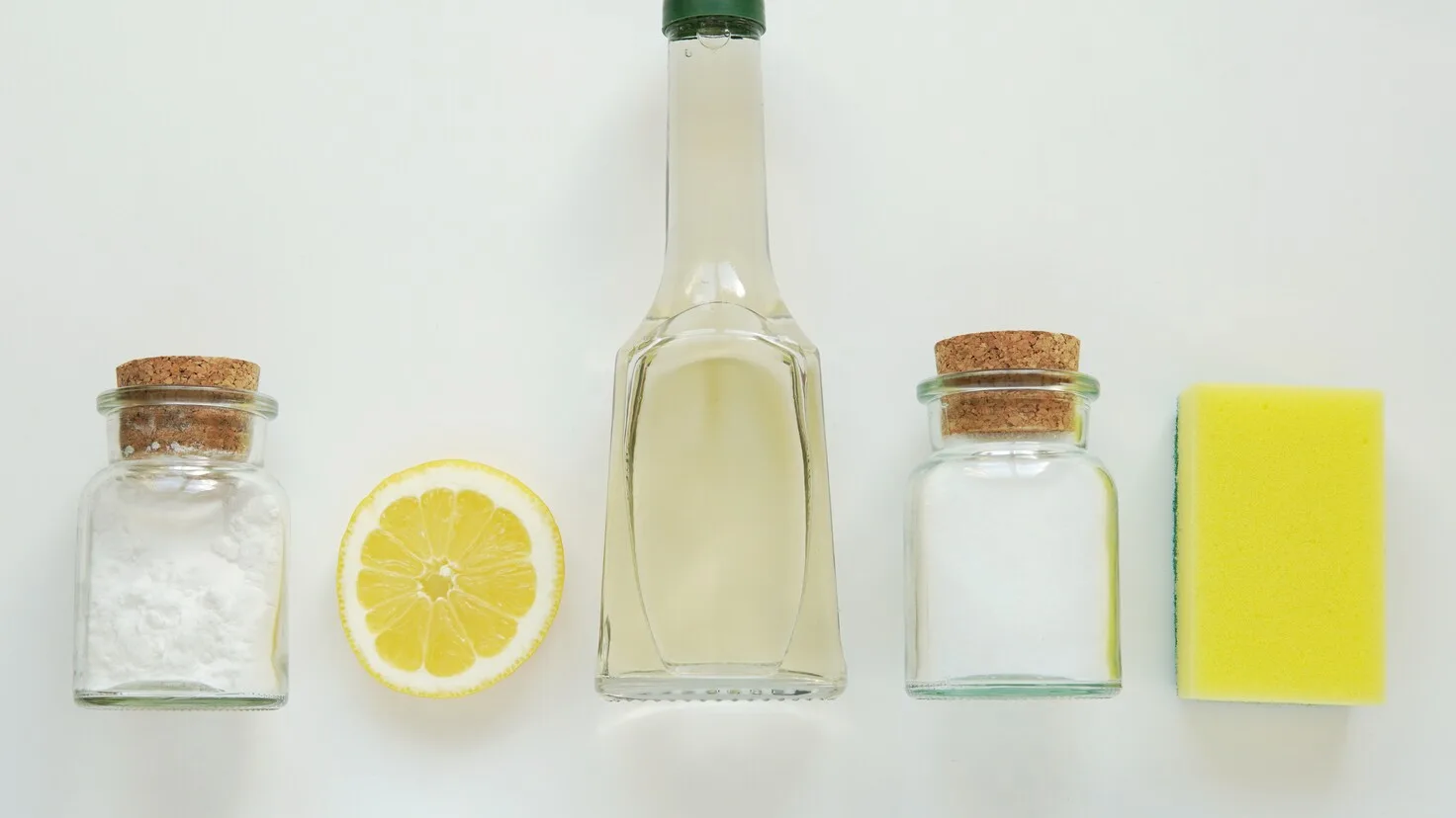 Produits ménagers maison alignés : bicarbonate, citron, vinaigre blanc, sel et éponge sur fond blanc