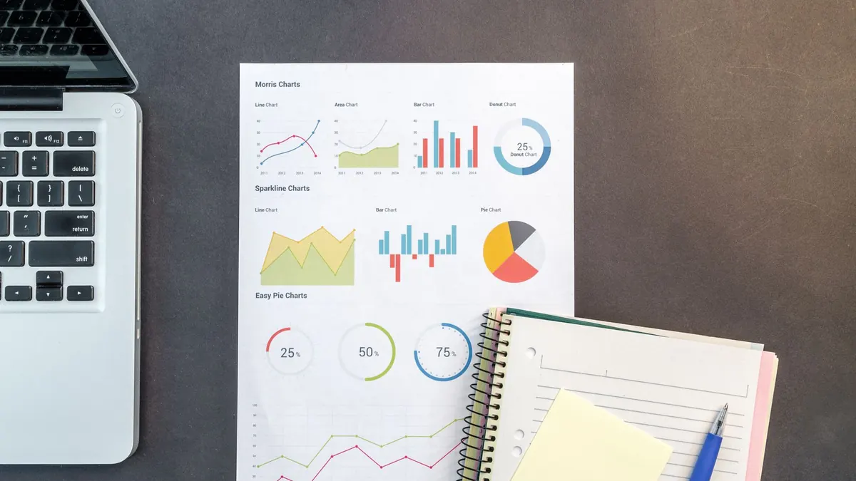 Bureau vu de dessus pour se constituer un patrimoine : feuille de graphiques financiers, carnet à spirale, stylo et ordinateur portable posés sur une table sombre